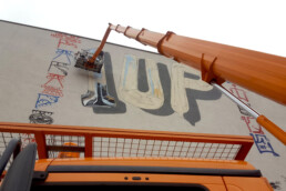 Hebebühne vor Fassadenbild: metallisch wirkende Bubble-Letter ‘1UP’