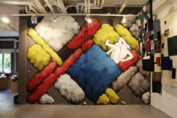 Ein lebendiges abstraktes Wandbild von Daan Botlek, das kräftige Formen und Farben zeigt, mit einem Galeriebereich darunter, der gerahmte Kunstwerke präsentiert.