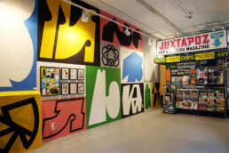 Ein lebendiges abstraktes Wandbild von Daan Botlek, das kräftige Formen und Farben zeigt, mit einem Galeriebereich darunter, der gerahmte Kunstwerke präsentiert.