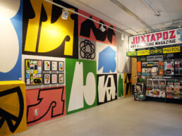 Ein lebendiges abstraktes Wandbild von Daan Botlek, das kräftige Formen und Farben zeigt, mit einem Galeriebereich darunter, der gerahmte Kunstwerke präsentiert.