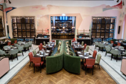 Blick in den Restaurantbereich des Fredericks Berlin mit gedeckten Tischen, zentraler Sitzbank und Bar im Hintergrund. Wandgestaltung von Johannes Mundinger.