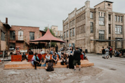 Besucherinnen und Besucher sitzen und stehen im Außenbereich des Monumenta Festivals Leipzig 2018 vor einer historischen Industriearchitektur.