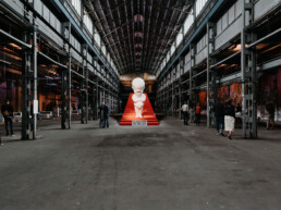Zentrale Skulptur auf roter Treppe in einer weitläufigen Industriehalle beim Monumenta Festival Leipzig 2018, Besucher im Raum verteilt.