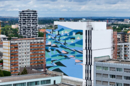 Weitblick auf das fertige Mural am MWG Forum in Magdeburg: grossflaechige, abstrakte Formen und die Schrift „FORUM MWG“ an der neuen Hauptzentrale.