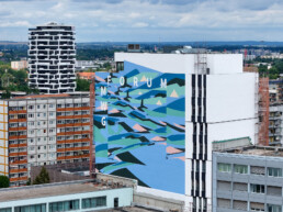 Weitblick auf das fertige Mural am MWG Forum in Magdeburg: grossflaechige, abstrakte Formen und die Schrift „FORUM MWG“ an der neuen Hauptzentrale.