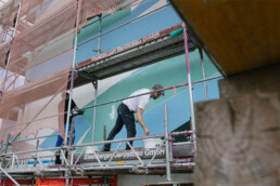 Kera und Team arbeiten auf dem Geruest und rollen Farbe auf die grossflaechigen, geometrischen Formen des Murals am MWG Forum in Magdeburg.