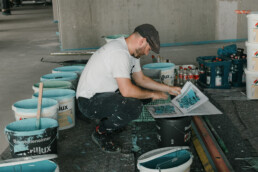 Vorbereitung der Mural-Produktion: Farbkuebel und Referenzmappe mit Entwurf, waehrend die Farben fuer Keras Fassadengestaltung am MWG Forum gemischt werden.