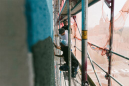 Das Team steht auf dem Geruest an der Fassade, im Hintergrund der Blick ueber Magdeburg waehrend der Mural-Produktion am MWG Forum.