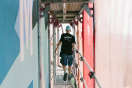 in Teammitglied im YAP-Shirt laeuft ueber den Geruestgang entlang der Fassade waehrend der Mural-Produktion von Kera am MWG Forum in Magdeburg.