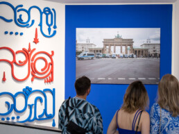 Besucher betrachten bei der Ausstellungseröffnung im URBAN NATION Museum eine typografische Wandinstallation von Paradox und eine Fotografie von Denis Cherim