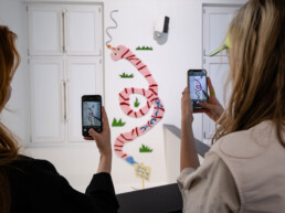 Besucherinnen fotografieren die Wandinstallation von HIN bei der Ausstellungseröffnung „Talking and other banana skins“ im URBAN NATION Museum Berlin.