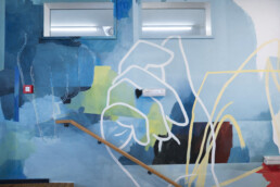 Abstrakte Wandgestaltung im Treppenaufgang des Vienna House Easy by Wyndham Berlin am Potsdamer Platz – Kunstwerk von Johannes Mundinger in Blau- und Grüntönen mit linearen Formen entlang des Geländers.