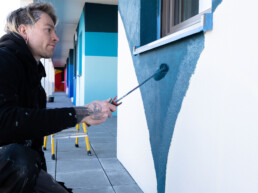 Assistent arbeitet an der Fassadengestaltung und trägt Farbe mit der Rolle auf – Produktion des Waterkant Murals.