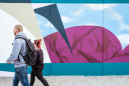 Passanten laufen an der 40 m Wandgestaltung für ANH Hausbesitz vorbei, abstrakte Formen in Türkis, Violett und Himmelblau.