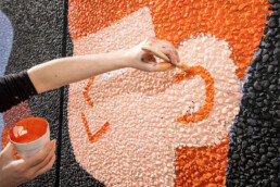 Brave Wall: Detailaufnahme – Hand mit Pinsel trägt orange Farbe auf strukturierte Wandfläche des Murals auf.