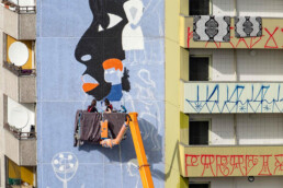 Brave Wall: Kate Voronina malt das Fassadenmural, Hubarbeitsbühne vor Wohnblock mit Balkonfassade.