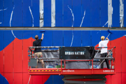 Farbauftrag an der Museumsfassade: Künstler rollen kräftiges Blau über orange-weiße Flächen an der URBAN NATION Fassade
