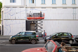 Vorzeichnung der Fassadengestaltung auf weißen Paneelen, zwei Künstler auf der Hebebühne an der URBAN NATION Museumsfassade