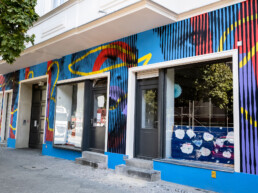 Straßenansicht einer Hausfassade mit farbigem Hundertzehn-Mural in Blau, Rot, Gelb und schwarzen Streifen