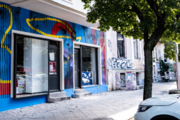 Seitliche Perspektive auf das Hundertzehn-Mural entlang der Straße, mit Baum im Vordergrund und angrenzender Hauswand