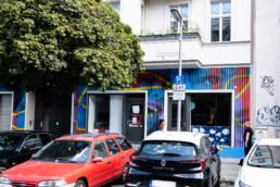 Breites Straßenmotiv mit parkenden Autos vor der bemalten Hundertzehn-Fassade und großem Schaufenster