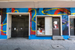 Hauseingang und Schaufenster vor Hundertzehn-Mural mit schwarzen Vertikalstreifen und gelben Linien auf blauem Hintergrund