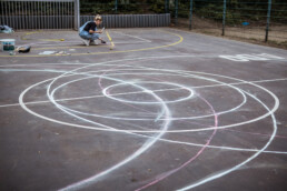 Kreideskizzen und Markierungen auf dem Basketballplatz als Vorzeichnung für die spätere Farbgestaltung, Person arbeitet im Hintergrund.