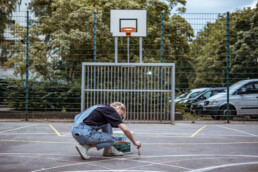 Person malt Linien auf dem Basketballplatz, Farbroller und Material stehen am Spielfeldrand, Korb und Zaun im Hintergrund.
