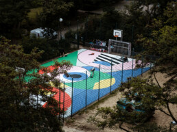 Luftaufnahme des Basketballplatzes während der Gestaltung: großflächige, farbige Formen und Arbeitsmaterialien im umzäunten Court.