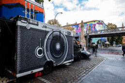 Photo-Booth-Truck mit Kamera-Grafik des Inside Out Projekts am Straßenrand, im Hintergrund Kreuzung und bemalte Fassade in Berlin.