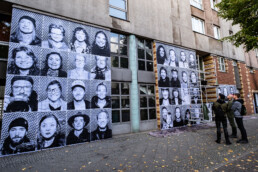 Großflächige Inside Out Porträtwände an einer Gebäudefassade, mehrere Personen betrachten die Montage vor Ort in Berlin.
