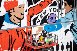 Lady Aiko arbeitet auf einer Hebebühne an der Urban Nation Museumsfassade; großformatige Malerei nach Motiven von Martha Cooper.