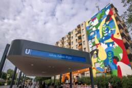 U-Bahnhof-Schild „Jakob-Kaiser-Platz“ im Vordergrund, im Hintergrund das fertige LandofJulia-Mural auf der Giebelfassade mit Hubarbeitsbühne.