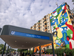 U-Bahnhof-Schild „Jakob-Kaiser-Platz“ im Vordergrund, im Hintergrund das fertige LandofJulia-Mural auf der Giebelfassade mit Hubarbeitsbühne.
