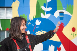 Portrait von LandofJulia vor dem Mural: erhobene Hand mit Farbresten, im Hintergrund grafische Pflanzen- und Farbformen.