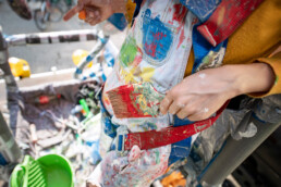 Nahaufnahme: Hand hält breiten Pinsel, Farbreste und Sprenkel auf Latzhose und Gurtzeug während der Mural-Produktion.