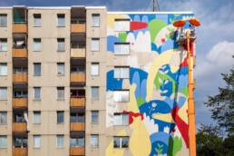 Weitblick auf Wohngebäude: große Giebelwand wird als Mural von LandofJulia gestaltet, Hubarbeitsbühne mit Schirmen am oberen Rand.