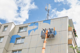 Zwei Personen auf einer Hubarbeitsbühne vor der Fassade, während die Vorzeichnung des Murals von LandofJulia aufgetragen wird.