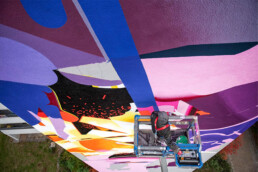 Detail der SatOne Fassadenmalerei: kräftige Farbflächen und Linien, Arbeitsszene auf der Bühne, Urban Nation