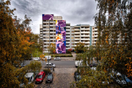 One Wall Mural von SatOne an einer Wohnhausfassade, Gesamtansicht zwischen Herbstbäumen, Urban Nation