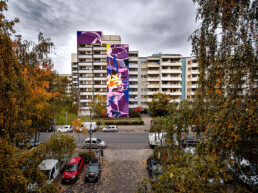 One Wall Mural von SatOne an einer Wohnhausfassade, Gesamtansicht zwischen Herbstbäumen, Urban Nation