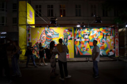 Abendstimmung vor einer großformatigen, bunt beleuchteten Wandgestaltung von Guapo Sapo beim UN-ARTIG Festival.