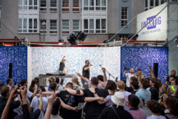 Publikum vor einer Bühne mit blau-weißer Line-Art-Kulisse von Leiti beim UN-ARTIG Festival, Urban Nation.