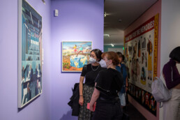 Besucher vor Kunstwerken an einer lila Wand in der Ausstellung „Talking and other Banana Skins“ im URBAN NATION Museum.