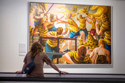 Besucher betrachtet ein großformatiges Gemälde mit gelb gekleideten Maskenfiguren in der Ausstellung „Talking and other Banana Skins“.