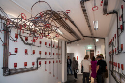 Rauminstallation mit Leitungen an Wänden und Decke sowie roten Griffen und Notruf-Elementen im URBAN NATION Museum.