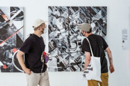 Zwei Besucher betrachten ein großformatiges Werk aus THE VERSUS PROJECT von Layer Cake in der URBAN NATION Ausstellung.