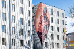 Großes Fassadenbild mit Porträt von Caro Pepe, Blick auf die Verbindung zwischen den Gebäudeteilen