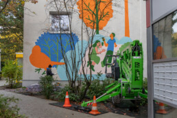 Arbeitsaufnahme der Vikunia Fassadengestaltung in der Mudrastraße für bbd mit grüner Hubarbeitsbühne vor dem fertigen Motiv.