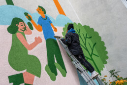 Vikunia arbeitet an der Fassadenmalerei in der Mudrastraße für bbd, Person auf Leiter ergänzt Figurenmotiv in Grün und Blau.